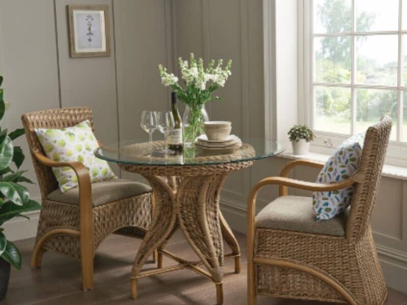 Two wicker chairs with patterned cushions face a round glass-topped table with wooden legs. On the table are white crockery, two wine glasses, a wine bottle, and a vase of white flowers. The scene is by a window with sunlight, wooden floor, and beige panelled walls.