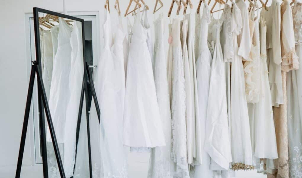 A row of white and cream dresses on wooden hangers is displayed on a clothing rail next to a large, black-framed standing mirror in a bright room with white walls and a door in the background. The scene is minimalistic and elegant.