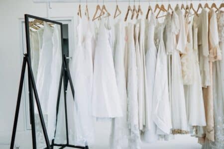A row of white and cream dresses on wooden hangers is displayed on a clothing rail next to a large, black-framed standing mirror in a bright room with white walls and a door in the background. The scene is minimalistic and elegant.