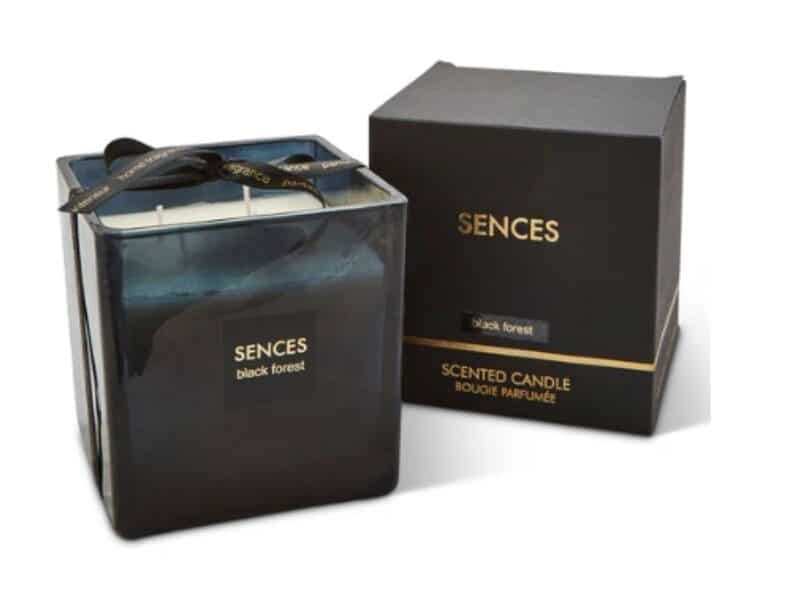 A black, square glass jar with a black ribbon and two white candle wicks labelled “SENCES black forest.” Next to it is a matching black box with gold text reading “SENCES black forest” and “SCENTED CANDLE BOUGIE PARFUMÉE”.