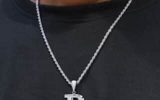 A person wearing a black shirt and a silver rope chain necklace with a large, diamond-encrusted letter "P" pendant hanging from it. The background is blurred and the focus is on the necklace and pendant.
