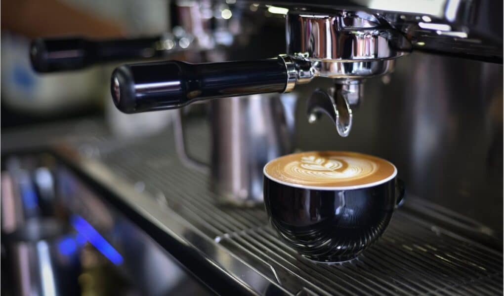 A black ceramic cup filled with coffee featuring intricate latte art sits on a metal drip tray of an espresso machine, with the portafilter and steam wand visible above, in a café or coffee shop setting.