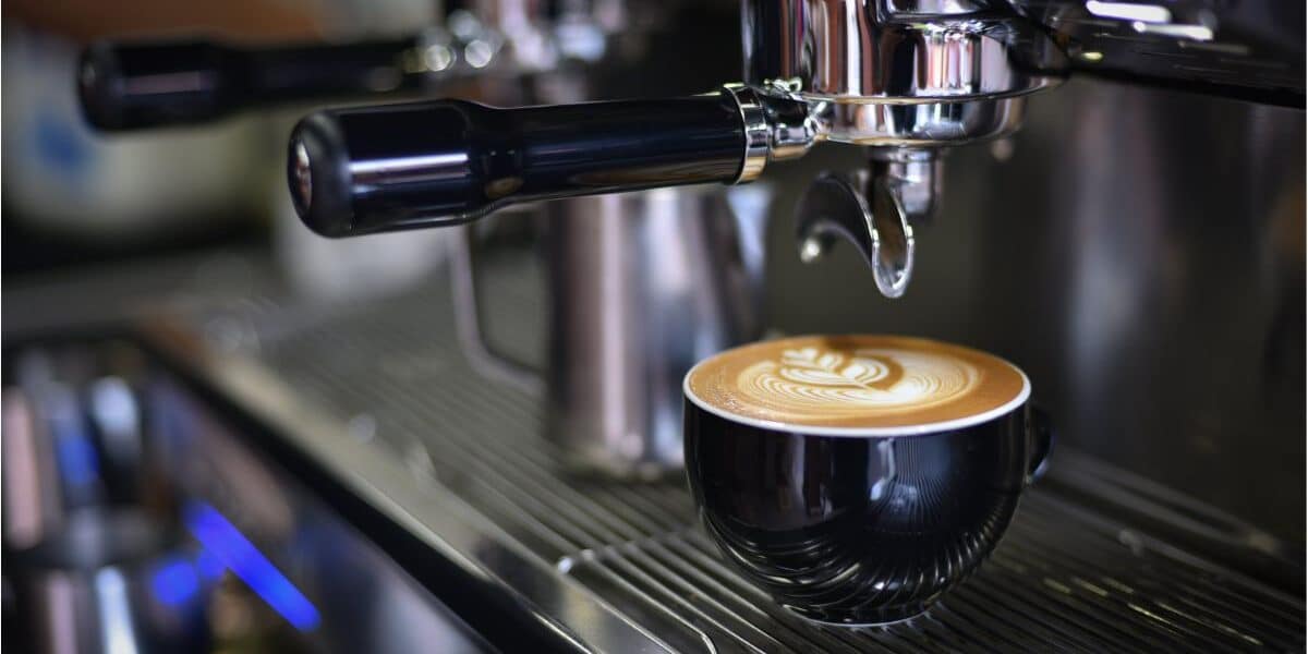 A black ceramic cup filled with coffee featuring intricate latte art sits on a metal drip tray of an espresso machine, with the portafilter and steam wand visible above, in a café or coffee shop setting.