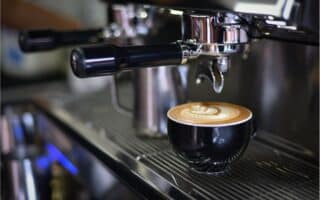 A black ceramic cup filled with coffee featuring intricate latte art sits on a metal drip tray of an espresso machine, with the portafilter and steam wand visible above, in a café or coffee shop setting.