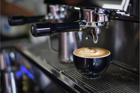 A black ceramic cup filled with coffee featuring intricate latte art sits on a metal drip tray of an espresso machine, with the portafilter and steam wand visible above, in a café or coffee shop setting.