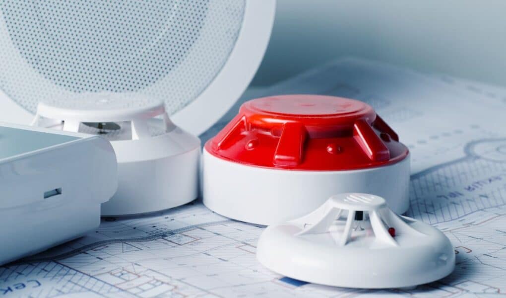 Close-up of smoke detectors and a red alarm siren placed on architectural blueprints, with a white round speaker or sensor in the background, suggesting planning or installation of a fire alarm or safety system in a building.
