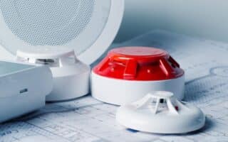 Close-up of smoke detectors and a red alarm siren placed on architectural blueprints, with a white round speaker or sensor in the background, suggesting planning or installation of a fire alarm or safety system in a building.