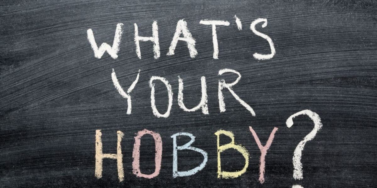 A blackboard with the words "WHAT'S YOUR HOBBY?" written in white chalk. The word "HOBBY" is written in all capital letters, each letter in a different colour: yellow, pink, blue, and peach. The background is dark and chalky.