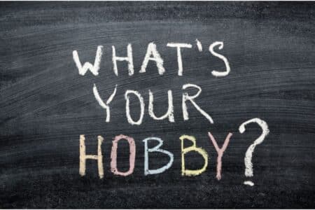 A blackboard with the words "WHAT'S YOUR HOBBY?" written in white chalk. The word "HOBBY" is written in all capital letters, each letter in a different colour: yellow, pink, blue, and peach. The background is dark and chalky.