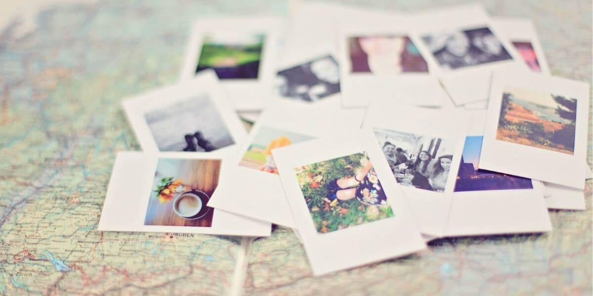 A pile of instant photo prints, some showing landscapes and people, lies scattered on a colourful, detailed map. The map’s roads and borders are visible beneath the photos, suggesting themes of travel and memories. The scene is softly focused and bright.