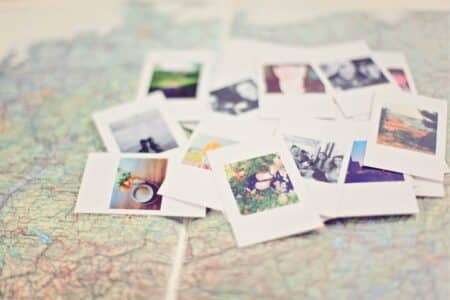 A pile of instant photo prints, some showing landscapes and people, lies scattered on a colourful, detailed map. The map’s roads and borders are visible beneath the photos, suggesting themes of travel and memories. The scene is softly focused and bright.