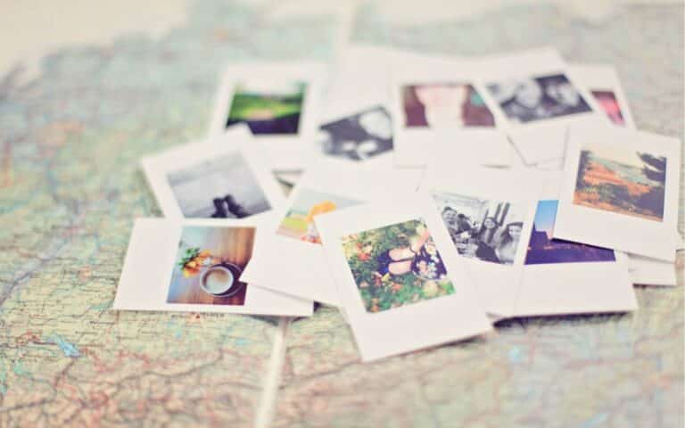 A pile of instant photo prints, some showing landscapes and people, lies scattered on a colourful, detailed map. The map’s roads and borders are visible beneath the photos, suggesting themes of travel and memories. The scene is softly focused and bright.