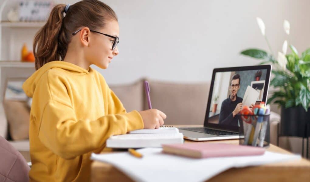 A girl in a yellow hoodie and glasses sits at a desk, writing in a notebook while looking at a laptop. On the screen, a man appears to be teaching or explaining something. The desk has books, pens, and pencils, with plants and a sofa in the background.
