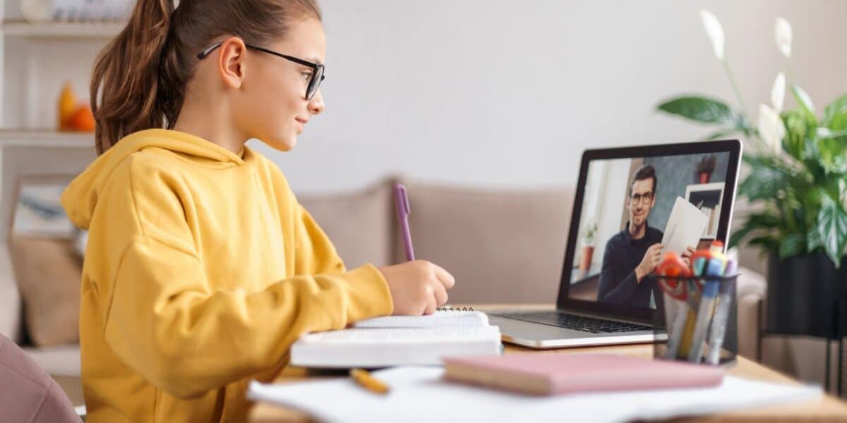 A girl in a yellow hoodie and glasses sits at a desk, writing in a notebook while looking at a laptop. On the screen, a man appears to be teaching or explaining something. The desk has books, pens, and pencils, with plants and a sofa in the background.