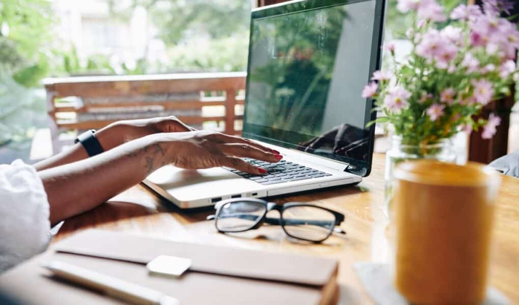 A person types on a laptop at a wooden table near a window. The table holds a closed notebook, spectacles, a pen, a glass of yellow drink on a coaster, and a vase of pink flowers. Sunlight and greenery are visible outside.