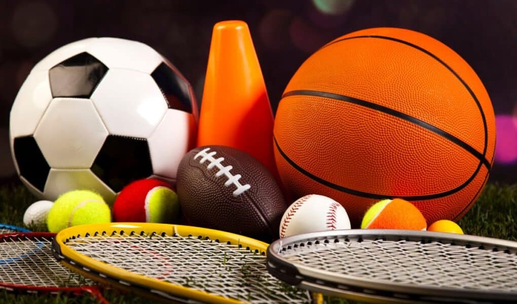 A collection of sports equipment, including a football, basketball, American football, several tennis balls, baseballs, an orange cone, and three tennis or badminton racquets, arranged on grass with a blurred dark background.