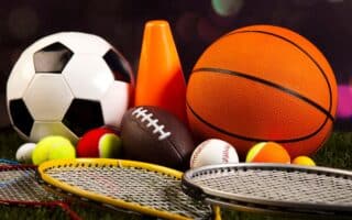 A collection of sports equipment, including a football, basketball, American football, several tennis balls, baseballs, an orange cone, and three tennis or badminton racquets, arranged on grass with a blurred dark background.