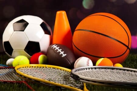 A collection of sports equipment, including a football, basketball, American football, several tennis balls, baseballs, an orange cone, and three tennis or badminton racquets, arranged on grass with a blurred dark background.