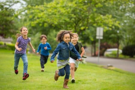 Four children run and smile on a grassy field in a park, with green trees and a road in the background. The kids wear casual clothes and appear to be playing together, enjoying a sunny day outdoors.