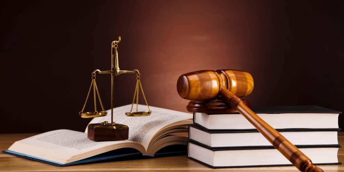A wooden judge’s gavel rests on two stacked books beside an open book. A brass set of balance scales stands between the books on a wooden table, with a dark brown background, symbolising law and justice.