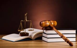 A wooden judge’s gavel rests on two stacked books beside an open book. A brass set of balance scales stands between the books on a wooden table, with a dark brown background, symbolising law and justice.