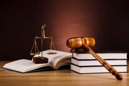 A wooden judge’s gavel rests on two stacked books beside an open book. A brass set of balance scales stands between the books on a wooden table, with a dark brown background, symbolising law and justice.