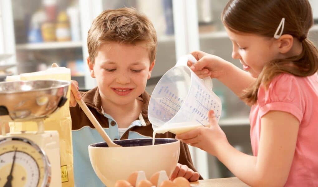 A young boy stirs a mixing bowl with a wooden spoon while a girl pours liquid from a measuring jug. Eggs, flour, and kitchen scales are on the table. Both children look focused and happy as they work together in a bright kitchen.