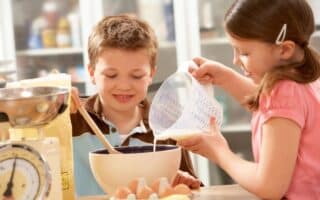 A young boy stirs a mixing bowl with a wooden spoon while a girl pours liquid from a measuring jug. Eggs, flour, and kitchen scales are on the table. Both children look focused and happy as they work together in a bright kitchen.