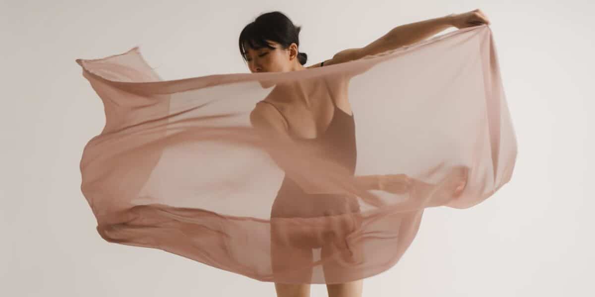 A woman in a leotard stands against a plain white background, holding and swirling a large, sheer, light pink fabric that billows around her, partially obscuring her figure. She gazes downwards, and her pose suggests movement or dance.