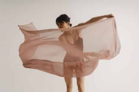 A woman in a leotard stands against a plain white background, holding and swirling a large, sheer, light pink fabric that billows around her, partially obscuring her figure. She gazes downwards, and her pose suggests movement or dance.