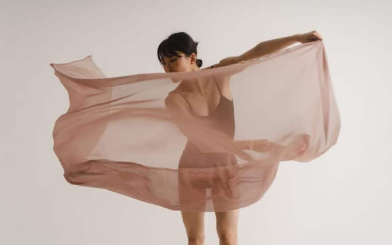 A woman in a leotard stands against a plain white background, holding and swirling a large, sheer, light pink fabric that billows around her, partially obscuring her figure. She gazes downwards, and her pose suggests movement or dance.