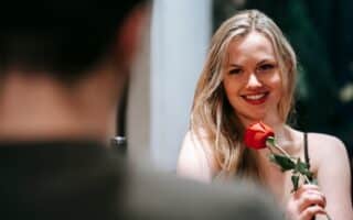 A smiling woman with long blonde hair holds a red rose whilst sitting at a table, looking at someone whose back is blurred in the foreground. She wears a black top and appears happy, in a warmly lit, possibly romantic setting.