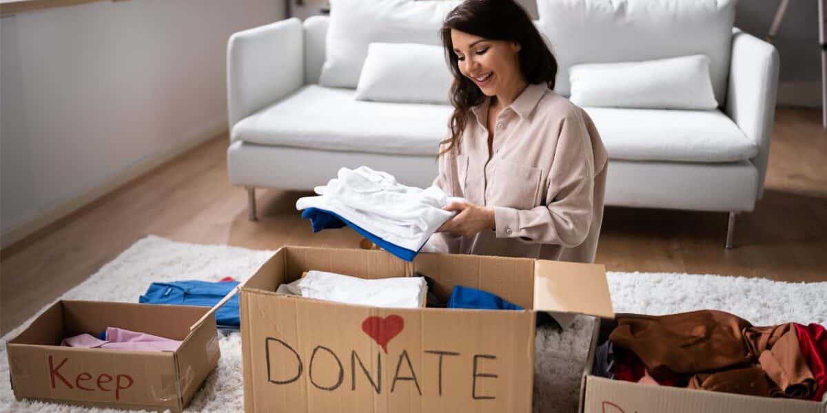 A woman sits on a carpeted floor in a lounge, smiling as she sorts clothes into three cardboard boxes labelled “Keep,” “Donate” with a red heart, and “Discard.” The room has a white sofa and bright natural light from large windows.