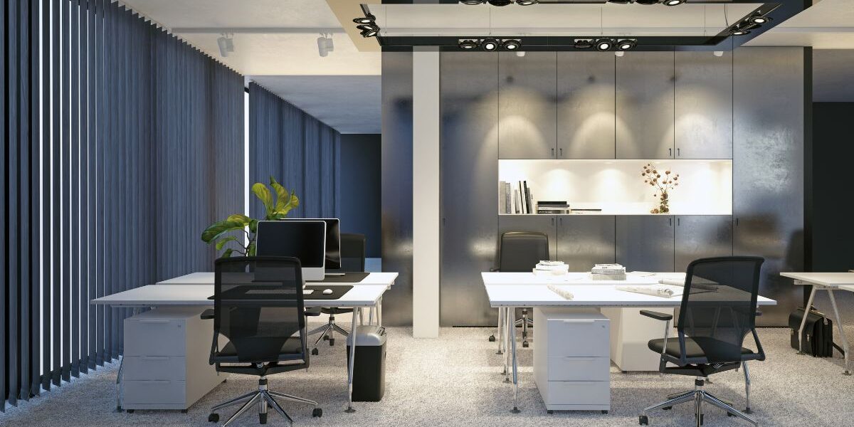 Modern office with grey carpeting, white desks, and black mesh chairs. Computers and phones sit on the desks. Vertical blinds cover tall windows on the left. A plant and shelves with decor are visible at the back, under ceiling light fittings.