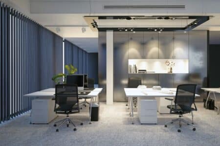 Modern office with grey carpeting, white desks, and black mesh chairs. Computers and phones sit on the desks. Vertical blinds cover tall windows on the left. A plant and shelves with decor are visible at the back, under ceiling light fittings.