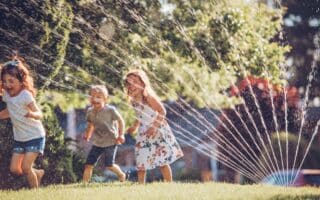 Three young children joyfully run through water spraying from a garden sprinkler on a sunny, grassy garden. Sunlight glistens on the water droplets, and the background is filled with green foliage, giving the scene a vibrant, playful summer feel.