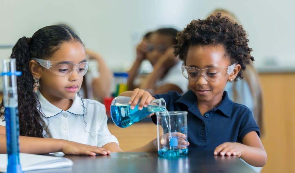 Two young girls wearing safety goggles work together in a science classroom. One girl pours blue liquid from a beaker into another beaker, while the other girl watches. Both are focused and seated at a black lab table with scientific equipment.
