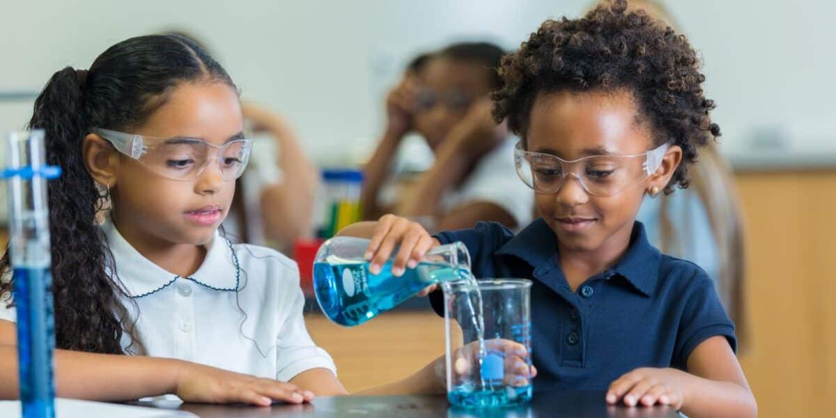 Two young girls wearing safety goggles work together in a science classroom. One girl pours blue liquid from a beaker into another beaker, while the other girl watches. Both are focused and seated at a black lab table with scientific equipment.