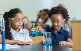 Two young girls wearing safety goggles work together in a science classroom. One girl pours blue liquid from a beaker into another beaker, while the other girl watches. Both are focused and seated at a black lab table with scientific equipment.