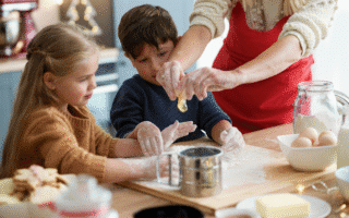 Two children with flour on their hands watch an adult squeeze an egg over a sieve on a messy kitchen worktop. The children look focused, surrounded by baking ingredients like butter, eggs, and flour. Biscuits and kitchen tools are also visible.