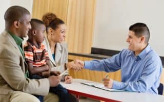 A man in a blue shirt smiles and shakes hands with a woman in a beige suit across a desk. Next to the woman sit a man in a tan suit and a young boy in a striped shirt; they all appear happy in a professional, welcoming setting.