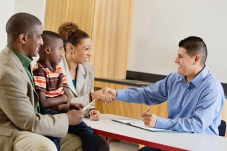 A man in a blue shirt smiles and shakes hands with a woman in a beige suit across a desk. Next to the woman sit a man in a tan suit and a young boy in a striped shirt; they all appear happy in a professional, welcoming setting.