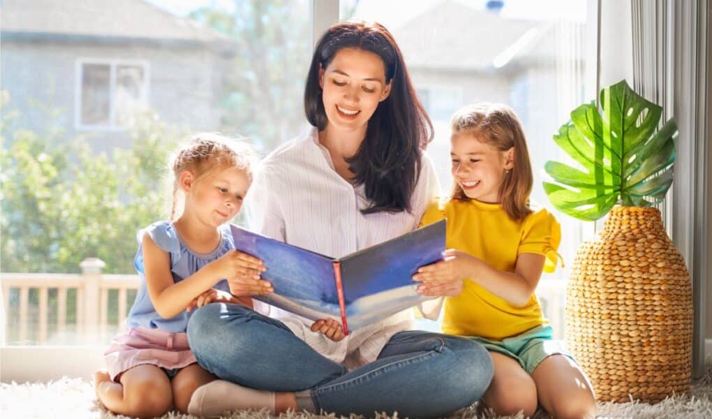 A woman sits cross-legged on a carpet, smiling while reading a large book to two young girls who sit on either side of her. Sunlight streams through large windows behind them. A woven basket and a green potted plant are on the right.