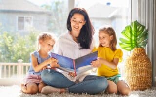 A woman sits cross-legged on a carpet, smiling while reading a large book to two young girls who sit on either side of her. Sunlight streams through large windows behind them. A woven basket and a green potted plant are on the right.