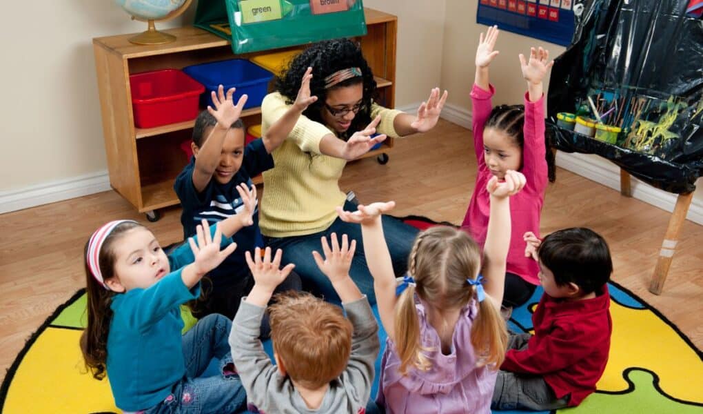 A teacher sits in a circle on a colourful rug with six young children, all raising their hands enthusiastically. The classroom has educational materials, including labelled bins, a globe, and a poster, creating a lively, engaging learning environment.