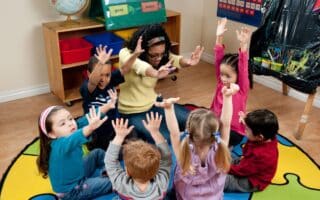 A teacher sits in a circle on a colourful rug with six young children, all raising their hands enthusiastically. The classroom has educational materials, including labelled bins, a globe, and a poster, creating a lively, engaging learning environment.