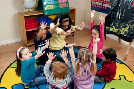 A teacher sits in a circle on a colourful rug with six young children, all raising their hands enthusiastically. The classroom has educational materials, including labelled bins, a globe, and a poster, creating a lively, engaging learning environment.