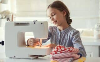 A young girl in a grey long-sleeve shirt uses a white sewing machine. She is smiling and focused on sewing yellow fabric. A stack of colourful, patterned cloth rests on the table beside her, and the room is brightly lit with natural light.