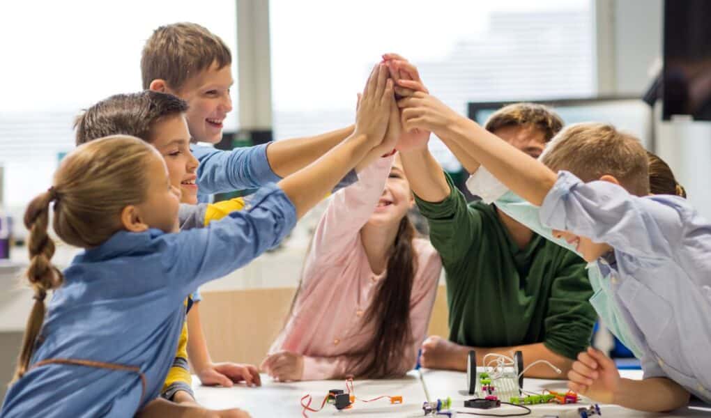 A group of six smiling children high-five each other over a table with electronics and wires, in a bright classroom. The kids appear happy and engaged, suggesting teamwork and collaboration on a science or STEM project.