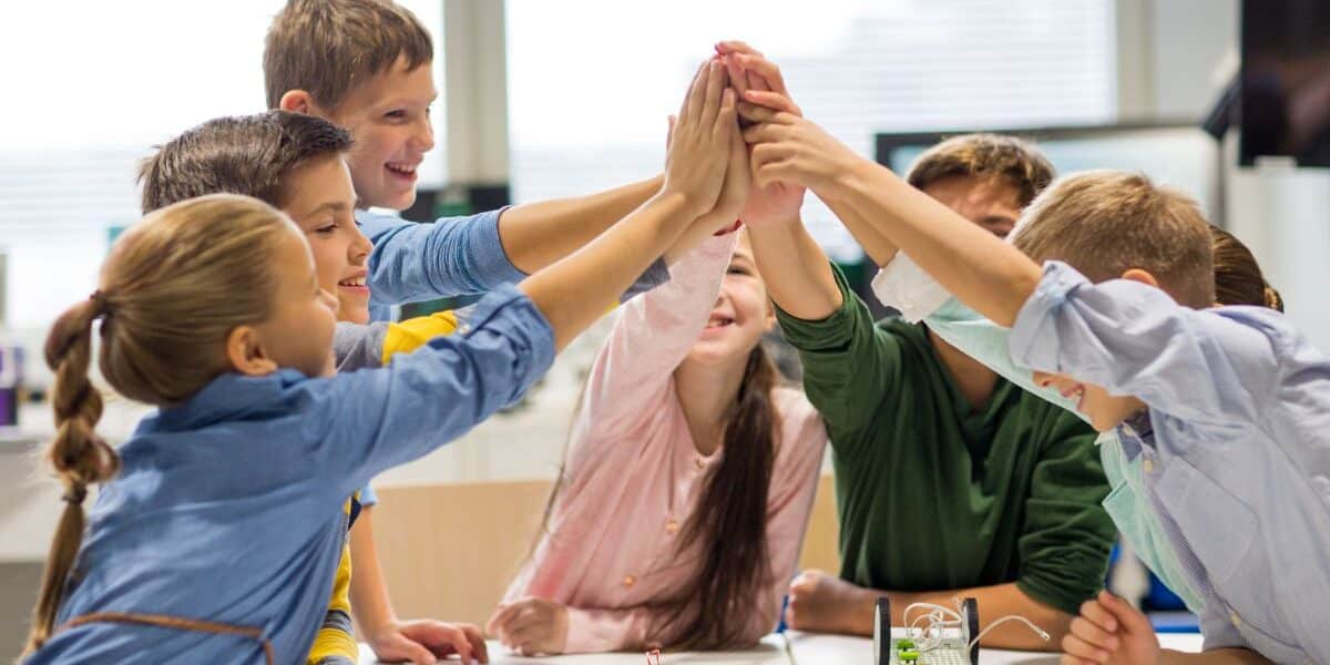 A group of six smiling children high-five each other over a table with electronics and wires, in a bright classroom. The kids appear happy and engaged, suggesting teamwork and collaboration on a science or STEM project.